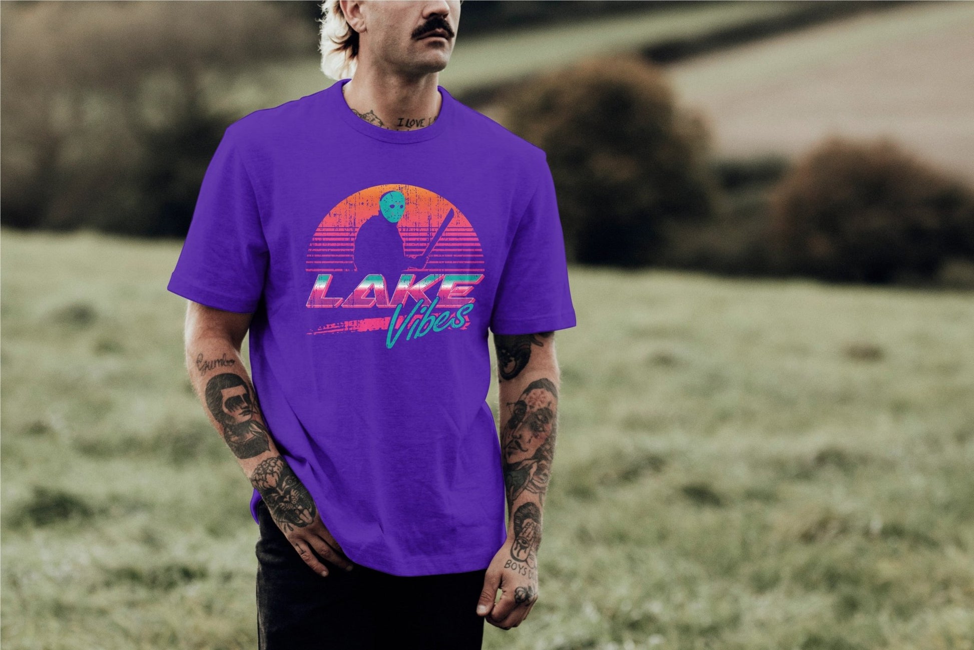 A man with tattoos and a mustache wearing a purple t-shirt with an orange sunset and Jason Vorhees in front of it and the words "Lake Vibes" underneath in a retro 80s style by Glow Bat Alternative Fashion