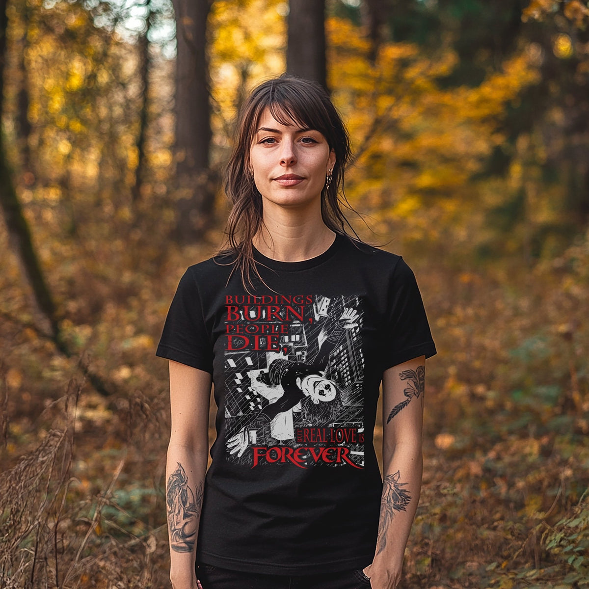 A woman with shoulder-length brown hair and arm tattoos, standing in a fall forest, wearing a black t-shirt from Glow Bat Alternative Fashion featuring the phrase "Buildings burn, people die, but real love is forever" from the 1994 goth horror film The Crow. along with an illustration featuring Eric Draven standing atop a cross on the steeple of a church, with his arms outstretched to the rainy sky with the city below.