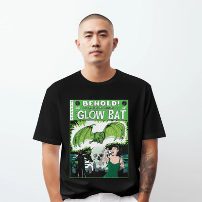A man with a shaved head wearing black t-shirt featuring an original green, white, and black illustration by Glow Bat Alternative Fashion of a dark figure summoning a large green bat towards an attractive dark-haired woman in a green dress. They are in a cemetery with a full moon with a skull in it. It is designed to look like an old horror comic like Tales From the Crypt.